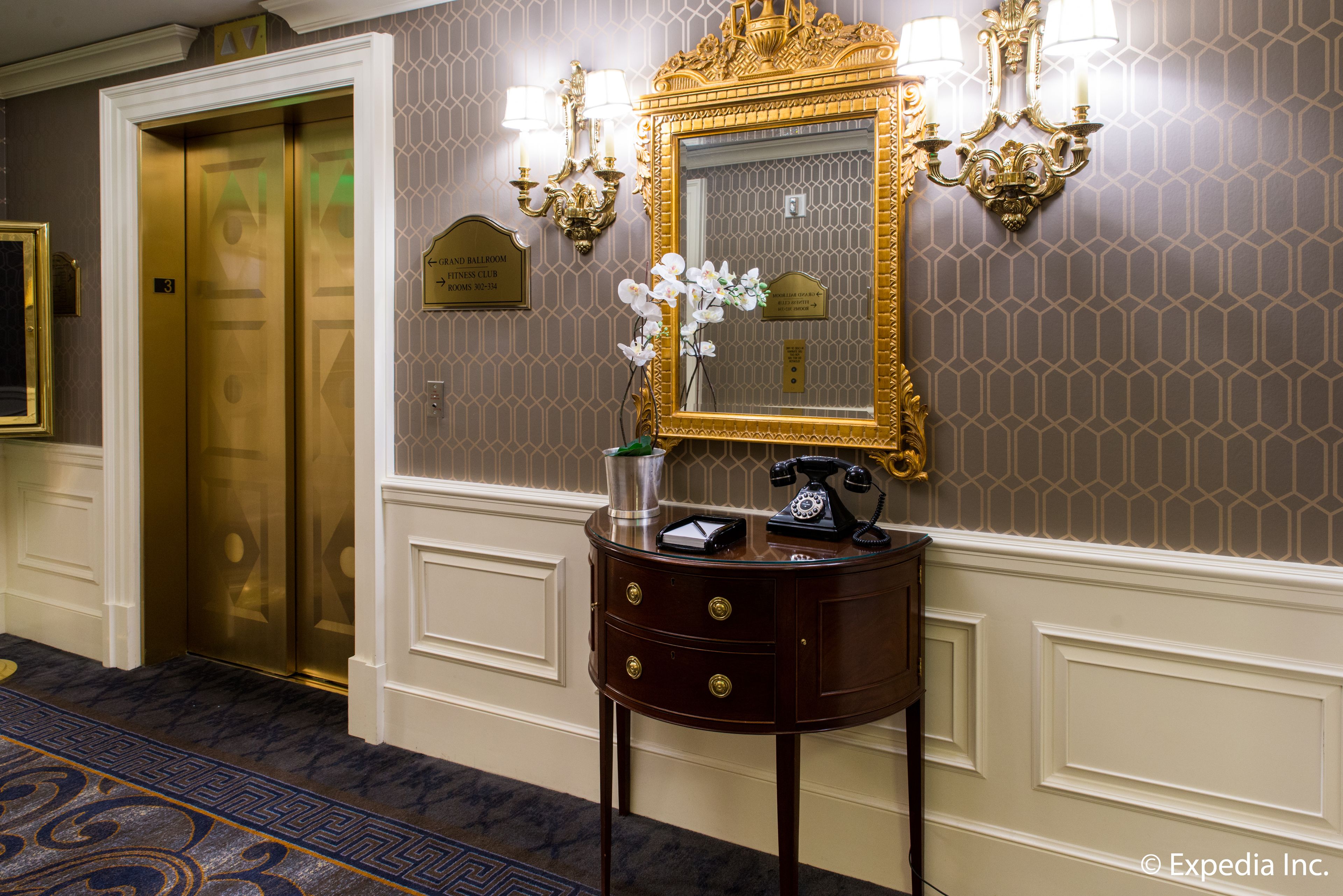 A hotel lobby with a gold elevator, a round wooden side table with a telephone, and a mirror with wall sconces.