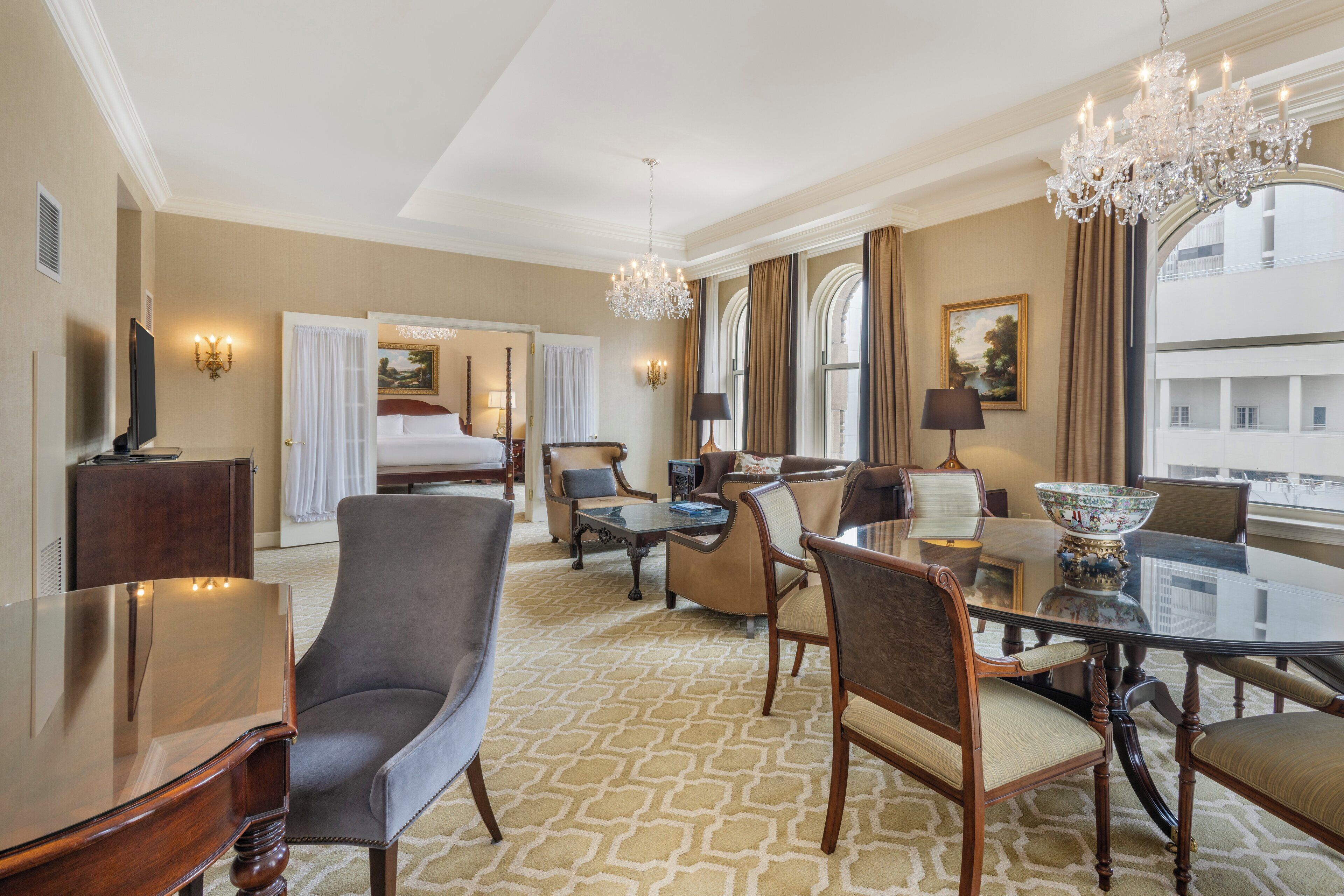 Habitación de hotel con una cama grande, una zona de estar con mesa y sillas de vidrio, televisión y candelabro.