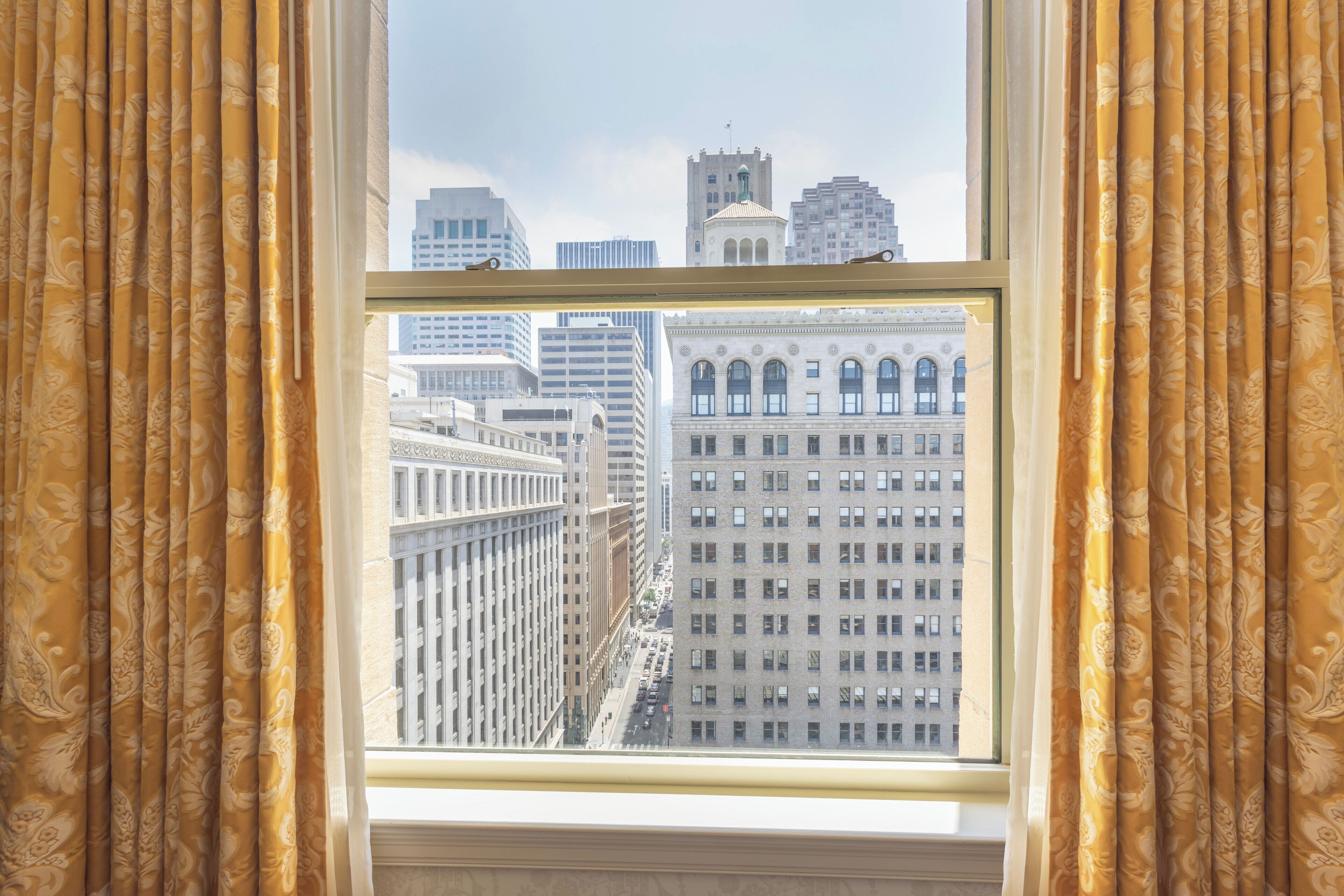 A window with ornate curtains, a view of a cityscape with tall buildings, and a clear sky.