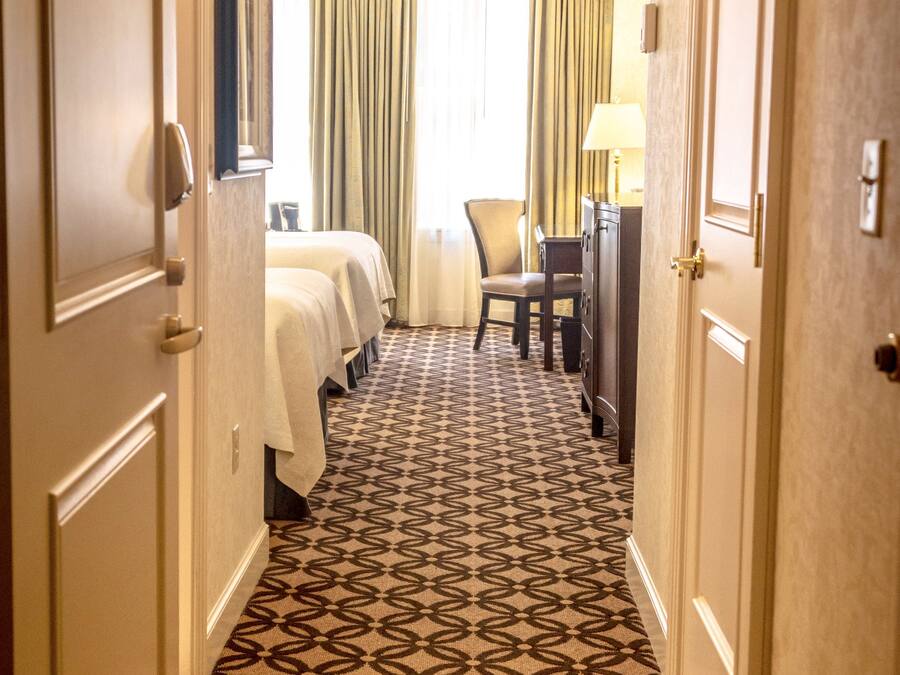 A hotel hallway with patterned carpet, a bed, a desk, and a chair.