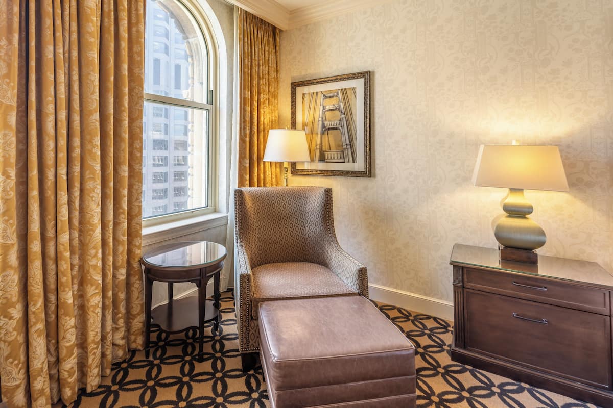 A hotel room with a patterned floor, a chair, a small table, a lamp, a cabinet, and a window with curtains.