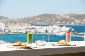 A table with breakfast items including toast, a green drink, a white bowl, and a pink drink, overlooking a coastal town. - Porto Mykonos Hotel (Mykonos)