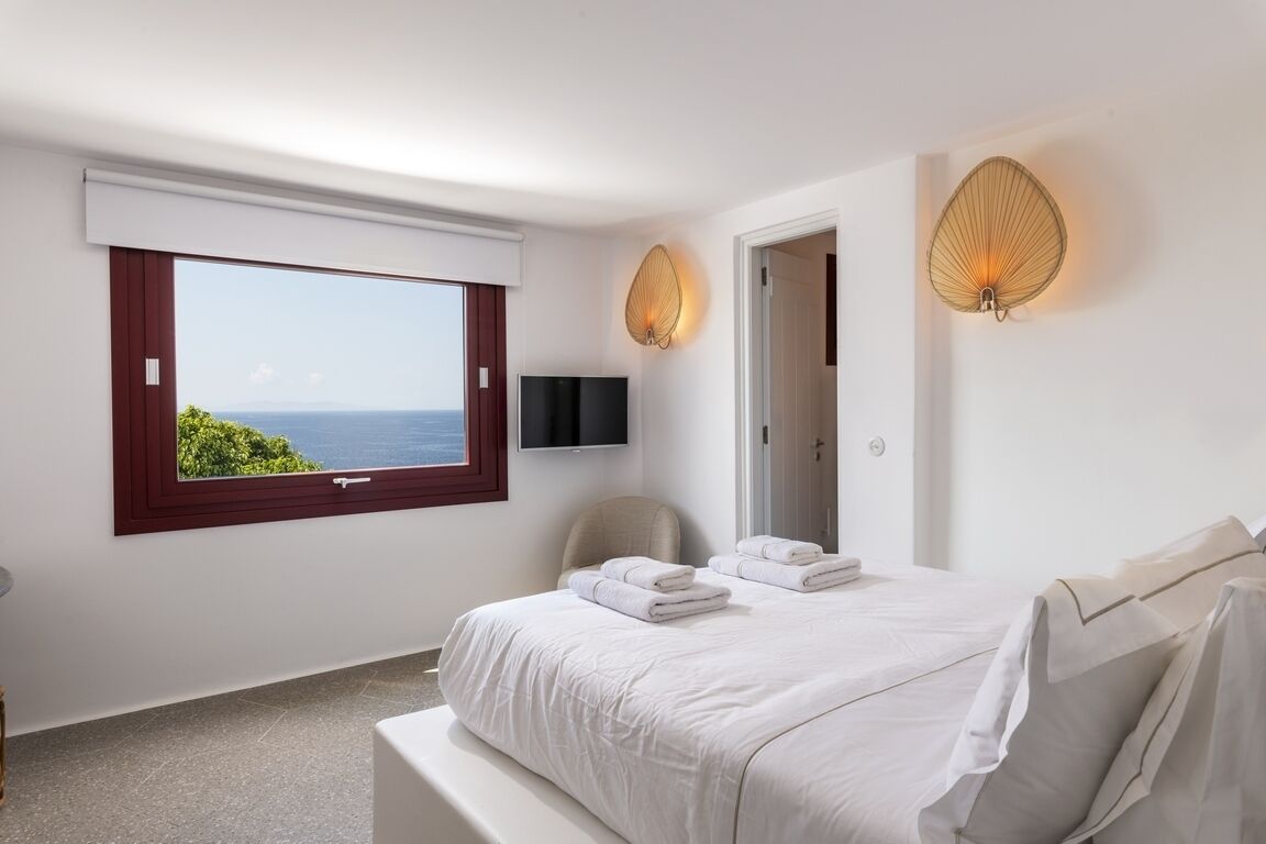 Un dormitorio con un ventanal grande, una cama con ropa de cama blanca, una silla y dos lámparas fijadas a la pared.