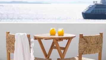 Una mesa redonda de madera con dos vasos de jugo de naranja, dos sillas de mimbre y un paño blanco cubriendo una silla. Se ve un barco grande al fondo.