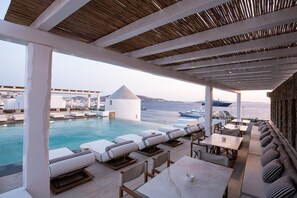 A poolside area with lounge chairs and tables, a view of the sea, and a white building in the background.