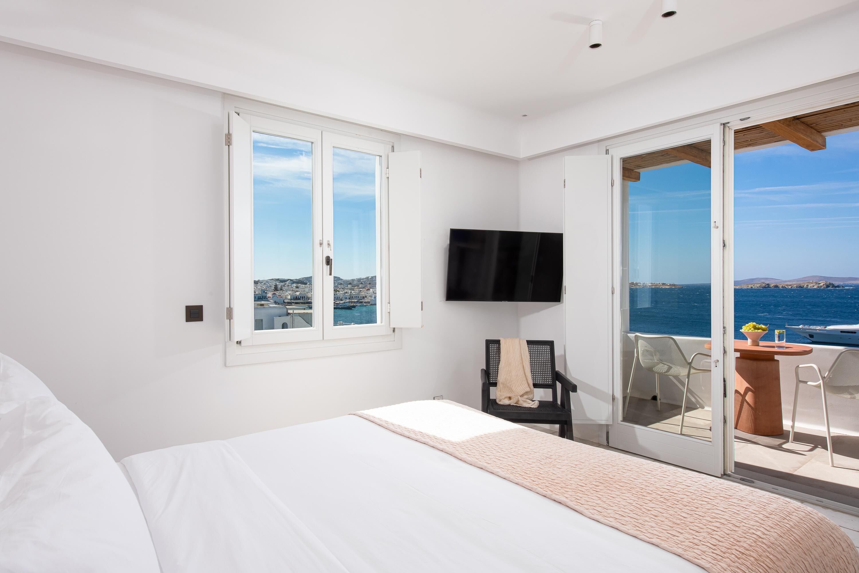 A bedroom with a bed, a TV, a chair, and a balcony with a view of the sea.