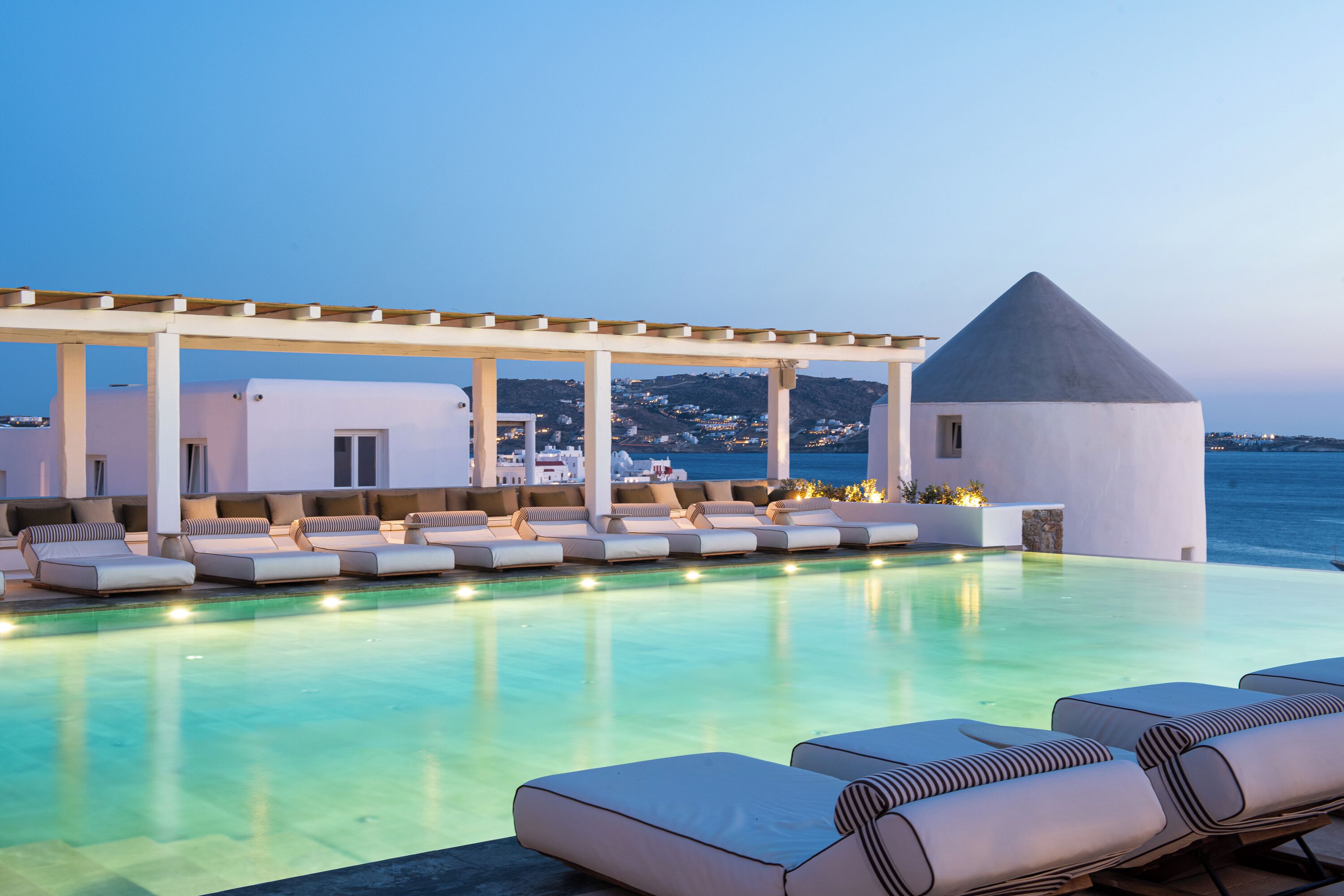 Une piscine à débordement située sur le toit, agrémentée de chaises longues et d’une pergola, offrant une vue sur une ville côtière.
