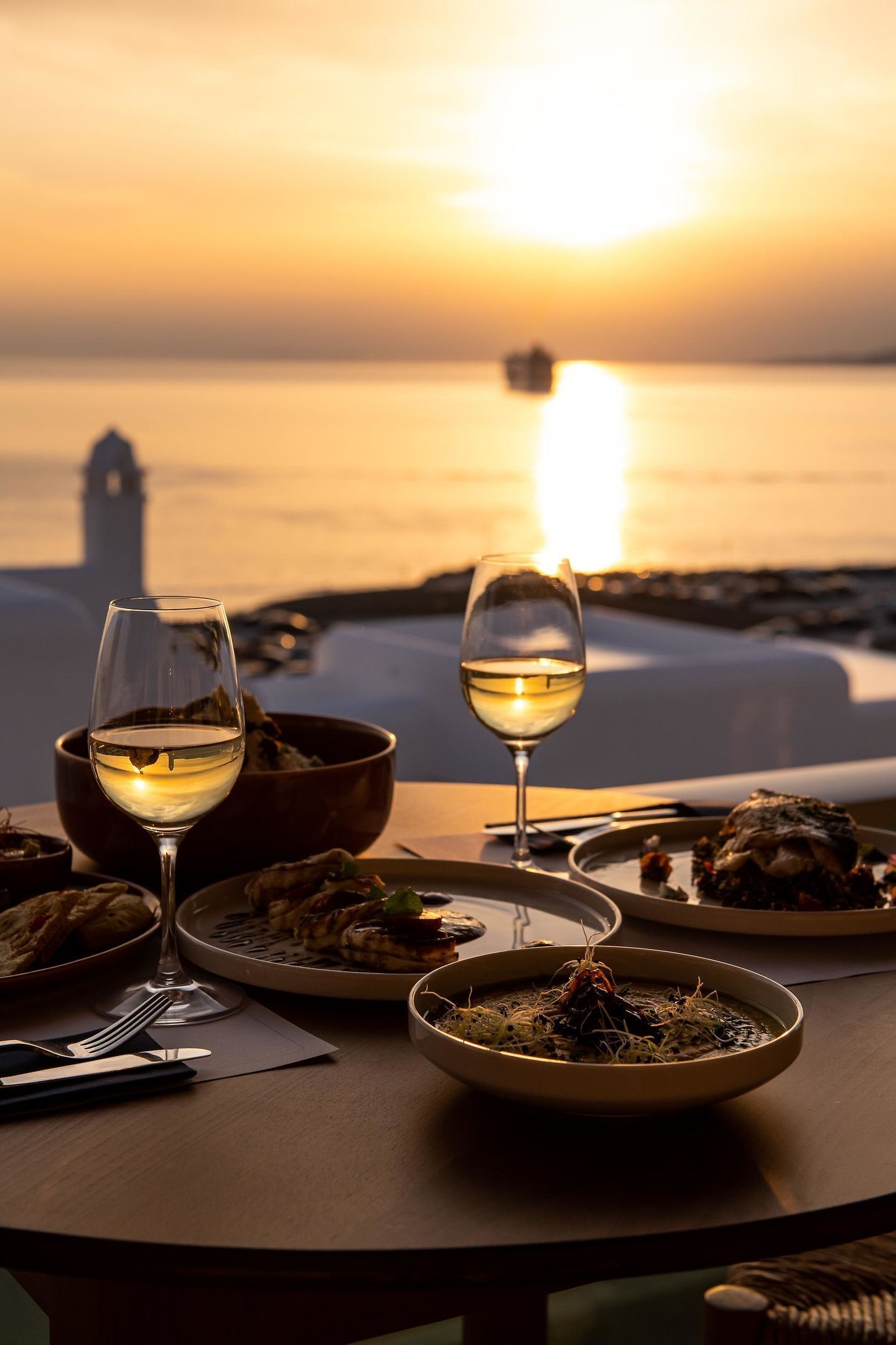 A table set for dinner with wine glasses, bowls of food, and a view of the sunset over the water.