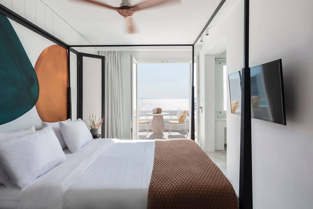 A modern hotel room with a large bed, a ceiling fan, a TV, and a balcony with a view of the sea.