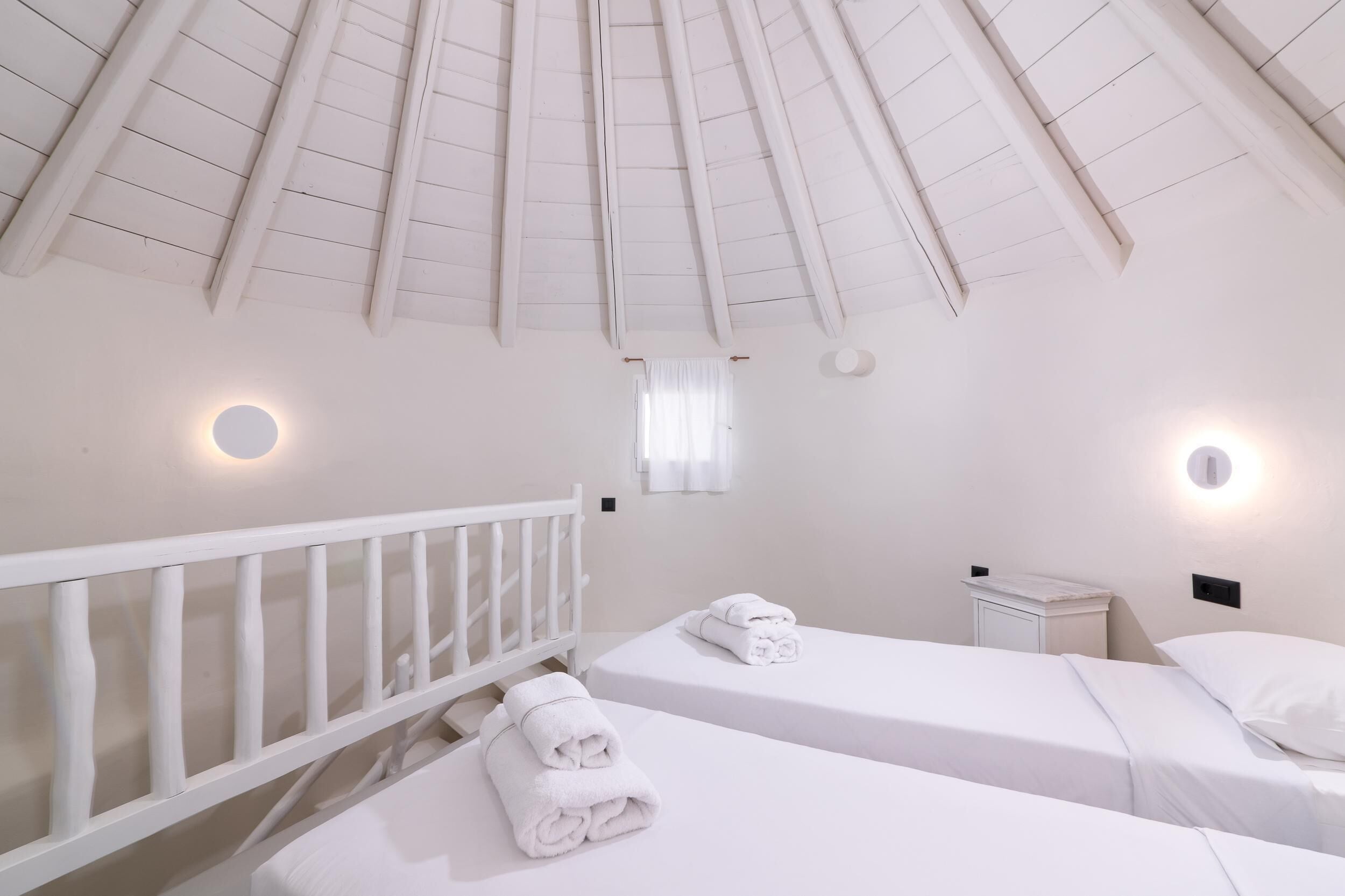 Una habitación con dos camas, una barandilla blanca, una mesita pequeña y un techo con vigas de madera.