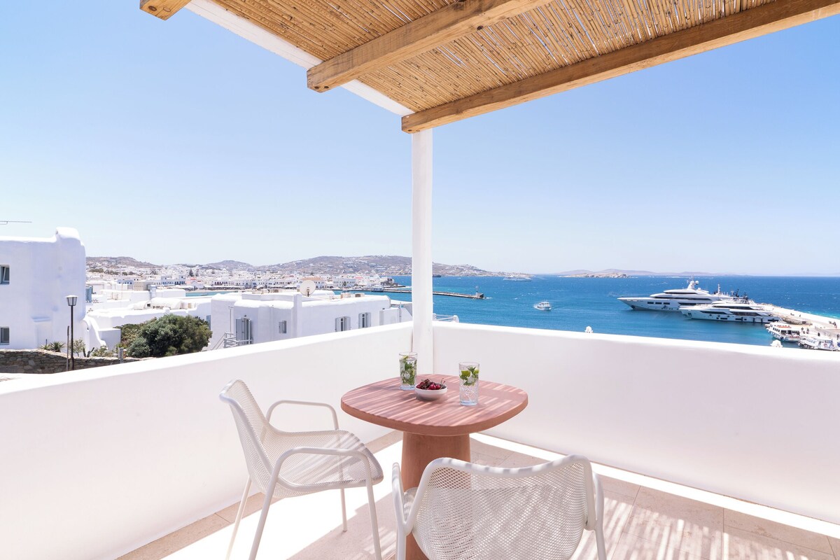 Ein Balkon mit rundem Tisch, zwei weißen Stühlen und Blick auf das Meer und Yachten.