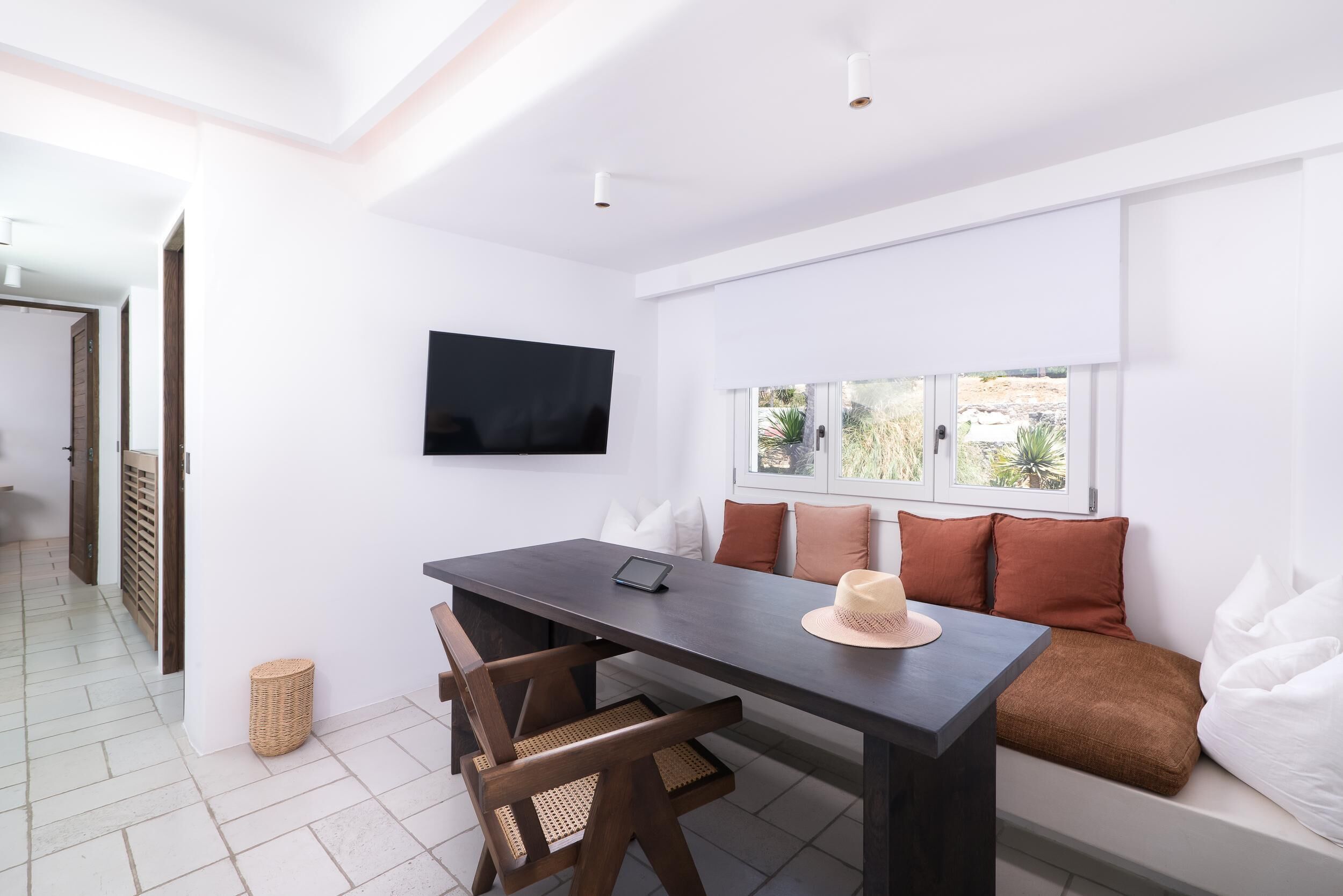 A modern room with a dark wooden table, two chairs, a TV mounted on the wall, a window with a view of greenery, and a small wicker basket on the floor.