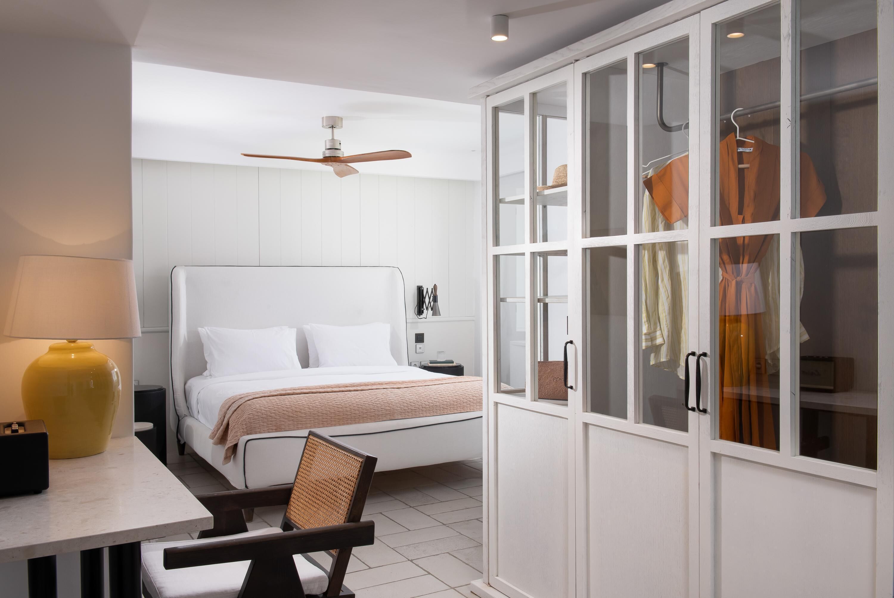 A bedroom with a bed, a chair, a desk with a lamp, a wardrobe with glass doors, and a ceiling fan.