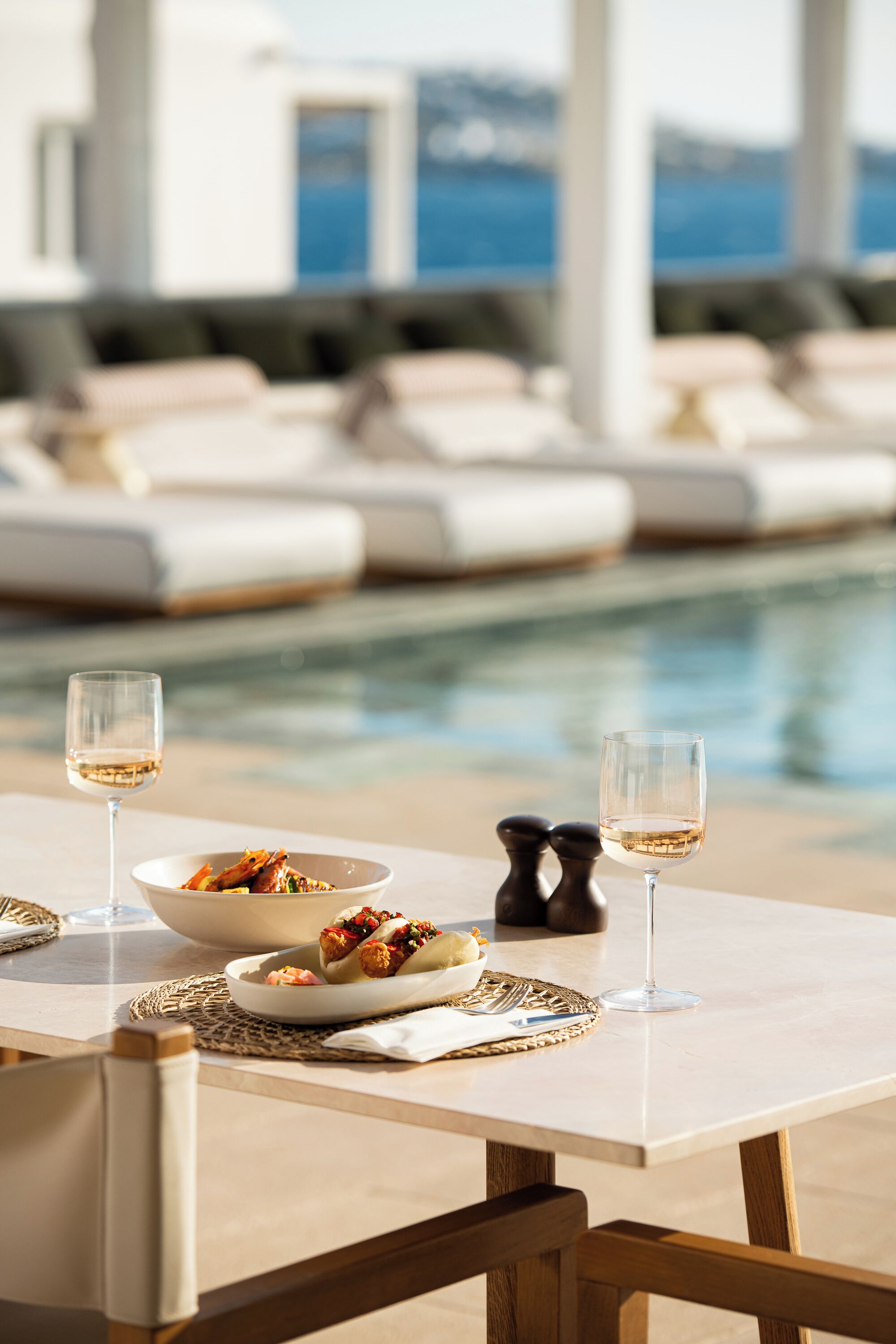 Une table à manger avec un saladier rempli de nourriture, deux verres à vin et des salières et poivrières. En arrière-plan, on aperçoit une piscine avec des chaises longues et une vue sur l’océan.