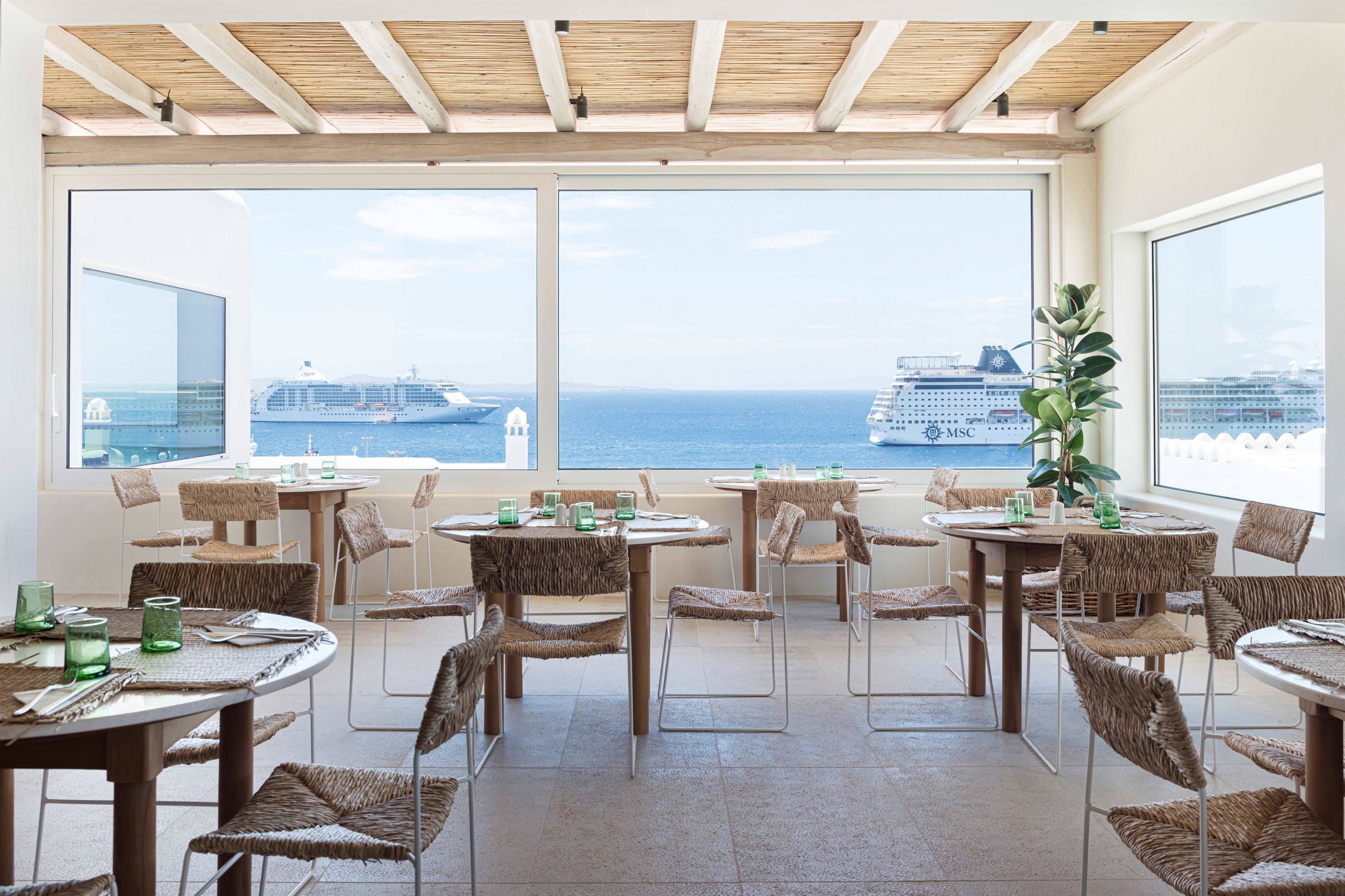 A dining area with wicker chairs and tables, large windows with a view of the sea and cruise ships.