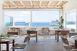 A dining area with wicker chairs and tables, large windows with a view of the sea and cruise ships. - Porto Mykonos Hotel (Mykonos)