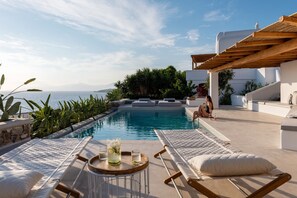 A poolside area with lounge chairs and a table with drinks, overlooking a body of water and greenery.