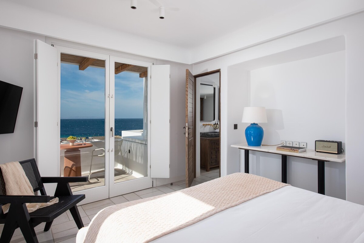 A bedroom with a bed, a desk with a lamp, a chair, a TV, and a sliding glass door leading to a balcony with a view of the sea.
