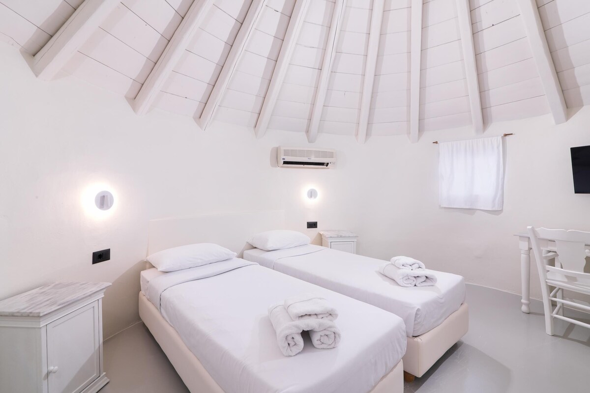 A hotel room with two beds, white bedding, a ceiling fan, an air conditioning unit, and a small bedside table.