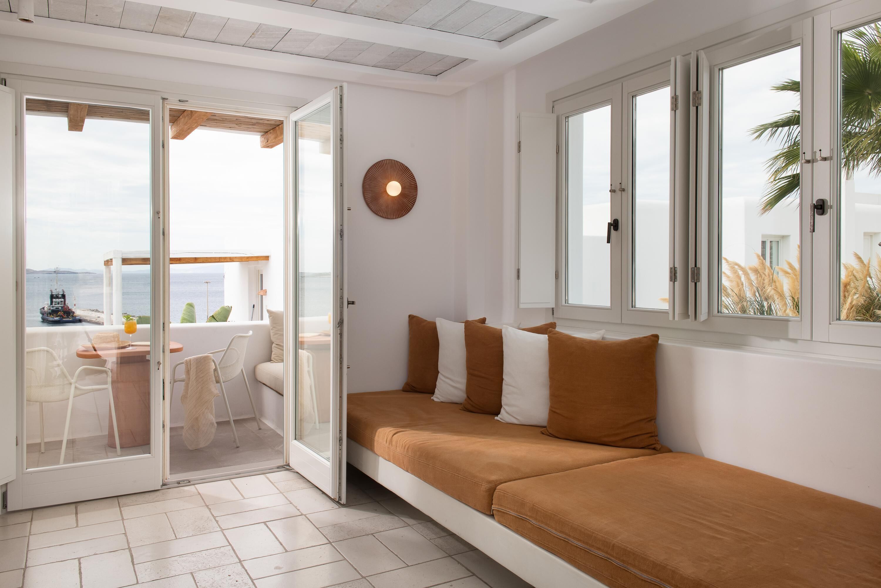 Quarto com porta de vidro deslizante para uma varanda, sofá com almofadas, luminária redonda fixada na parede e vista para o mar.