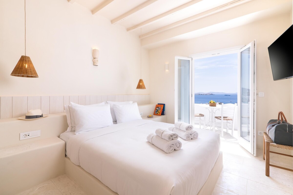 A bedroom with a large bed, white bedding, a bedside table, a lamp, and a view of the sea through an open door.