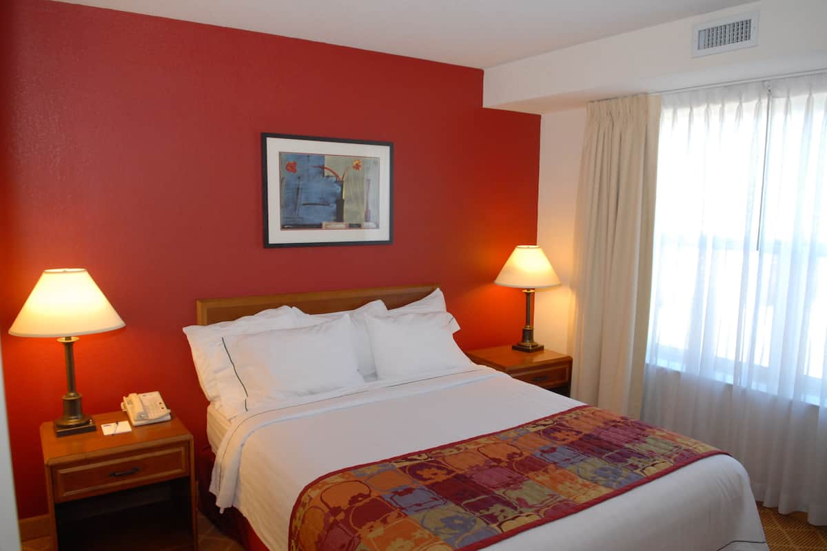A hotel room with a bed, two bedside tables with lamps, a framed picture on the wall, and a window with curtains.