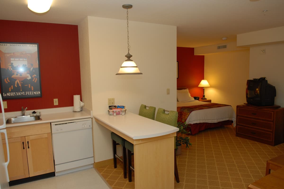 A compact hotel room with a kitchenette, a bed, a TV, and a bedside lamp.