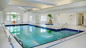 Una amplia piscina interior con agua cristalina y de color azul, rodeada de paredes blancas y suelo de baldosas. Se observan plantas en macetas y un reloj en la pared.