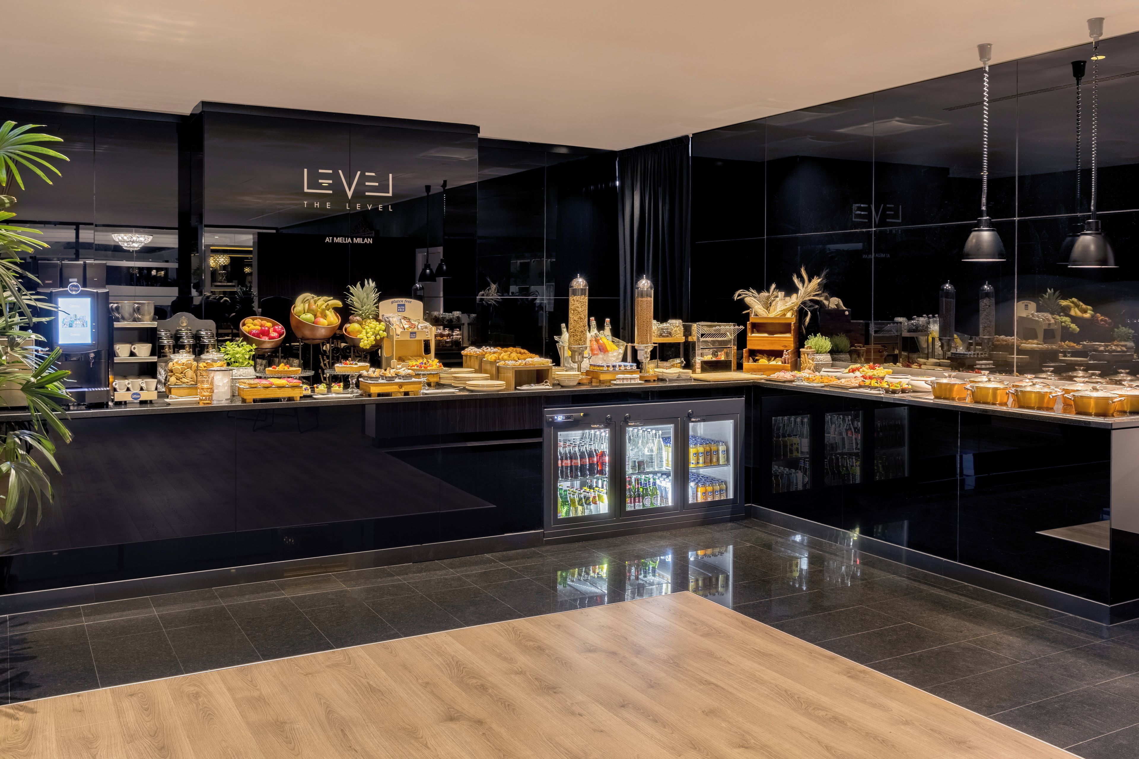 Ett modernt barområde med en färgskala i svart och trä, med en kaffemaskin, ett varierat utbud av mat och dryck samt ett kylskåp för att visa upp produkterna.