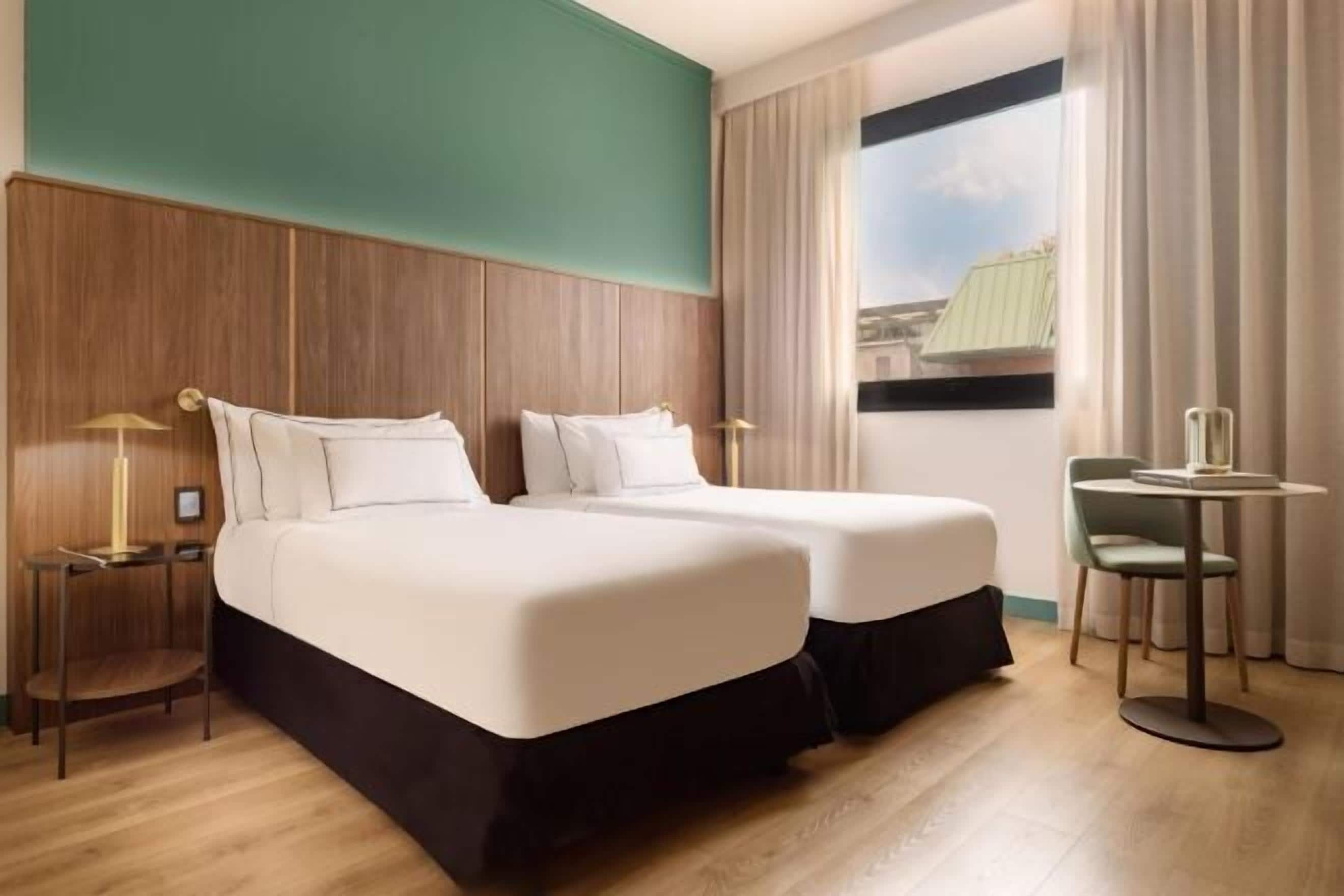 A hotel room with two beds, a small table, and a view of the city.