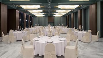 A large, round dining room with multiple tables set for a formal event, featuring chandeliers and tall, narrow windows.