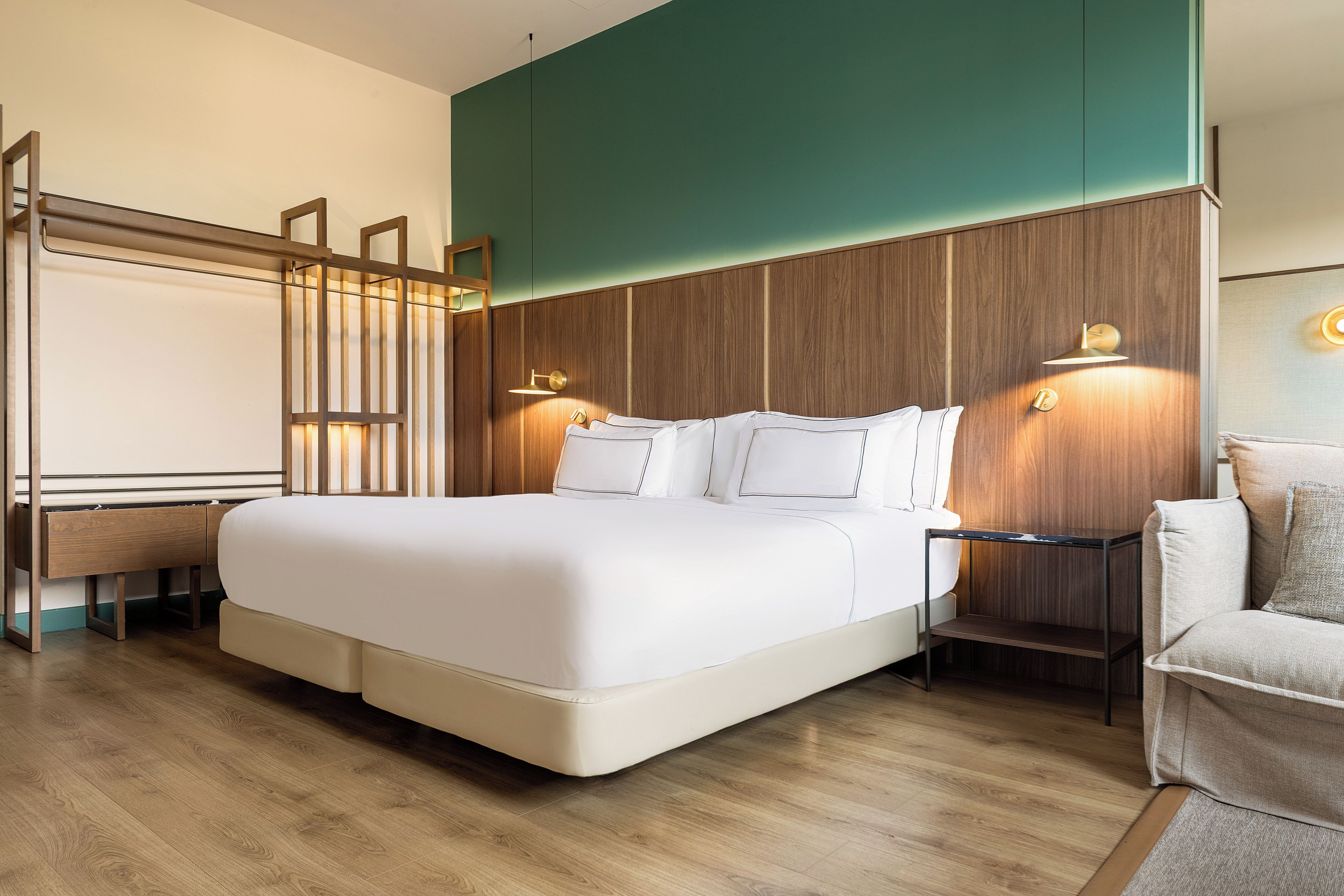 A modern hotel room with a large bed, wooden paneling on the walls, a bedside table with a lamp, and a seating area with a sofa.