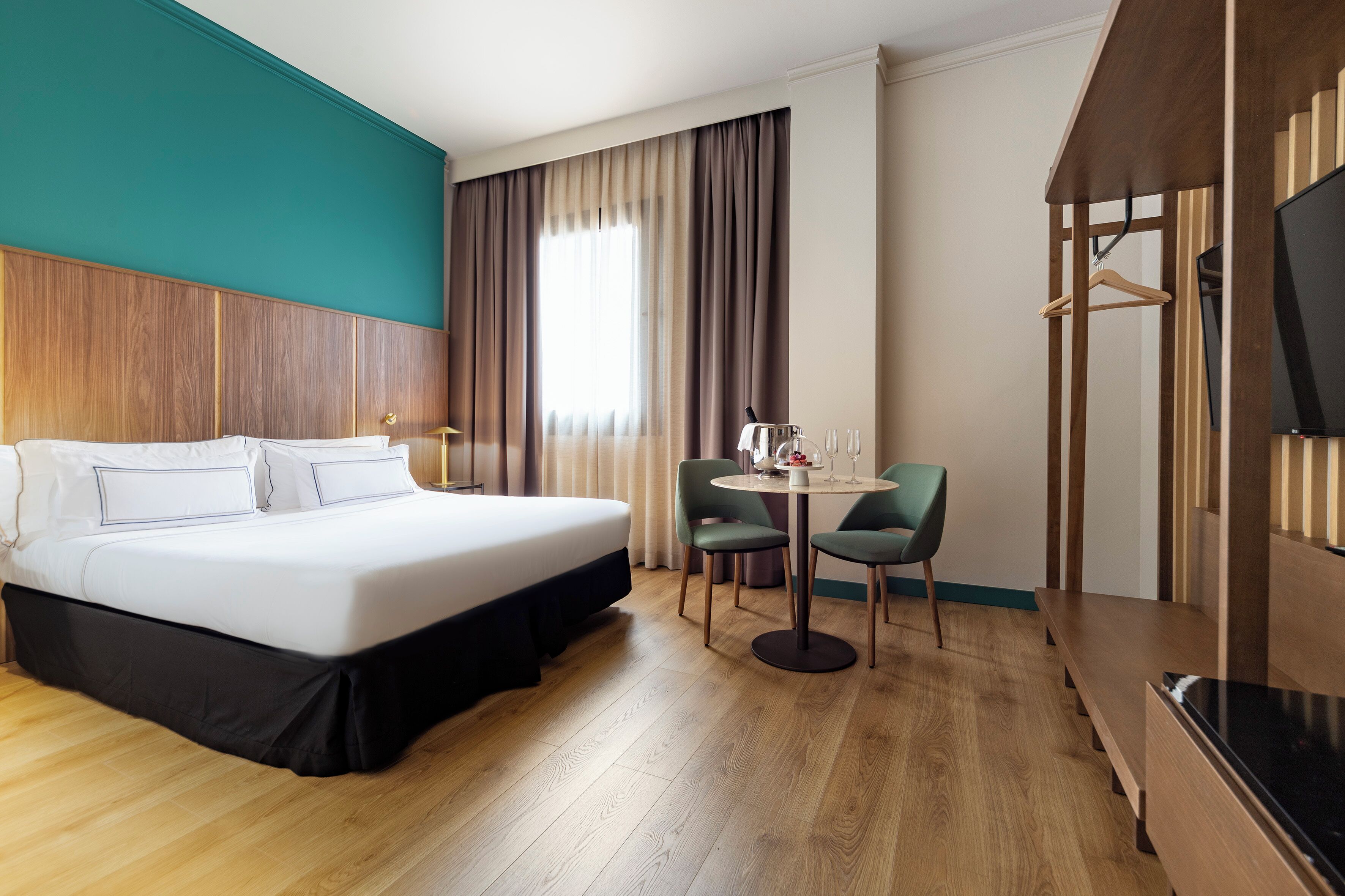 Habitación de hotel con cama, escritorio con sillas, televisión y ventana con cortinas.