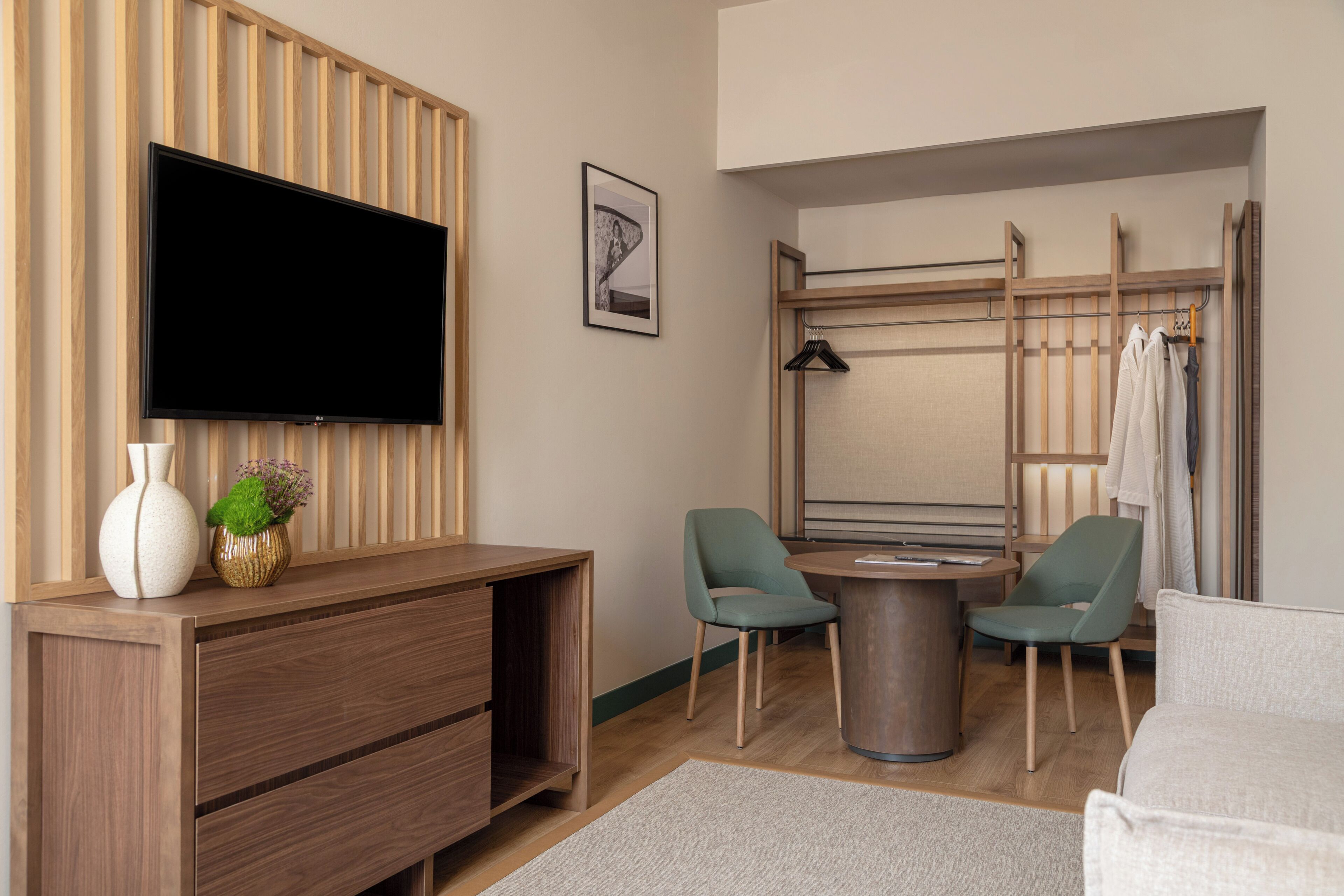 A modern hotel room with a flat-screen TV mounted on the wall, a wooden console table, two green chairs, a round coffee table, and a bed.