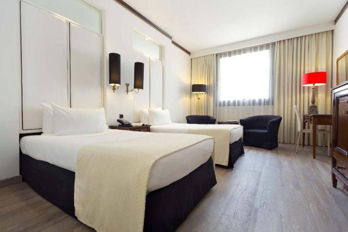A hotel room with two beds, a desk, a chair, a lamp, and a large window with curtains.
