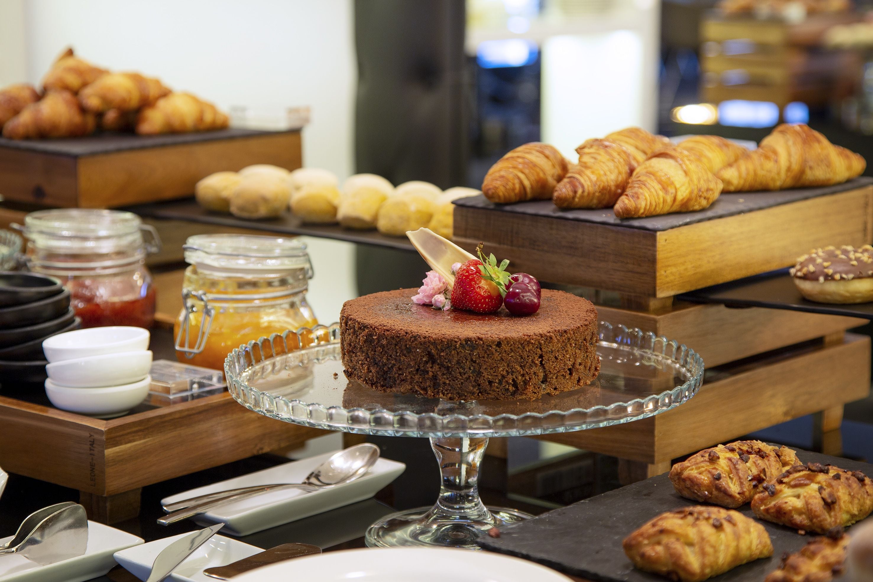 A buffet with various baked goods, including croissants and pastries, and a chocolate cake with strawberries on a glass stand.