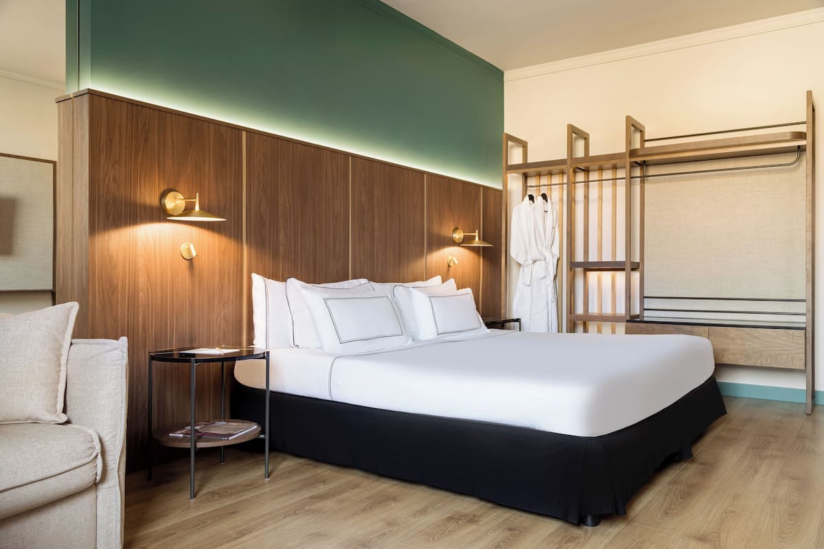 Une chambre d’hôtel moderne avec un grand lit, une tête de lit en bois, une table de chevet et un coin salon avec un fauteuil.