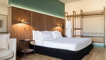 A modern hotel room with a large bed, a wooden headboard, a nightstand, and a seating area with a chair.