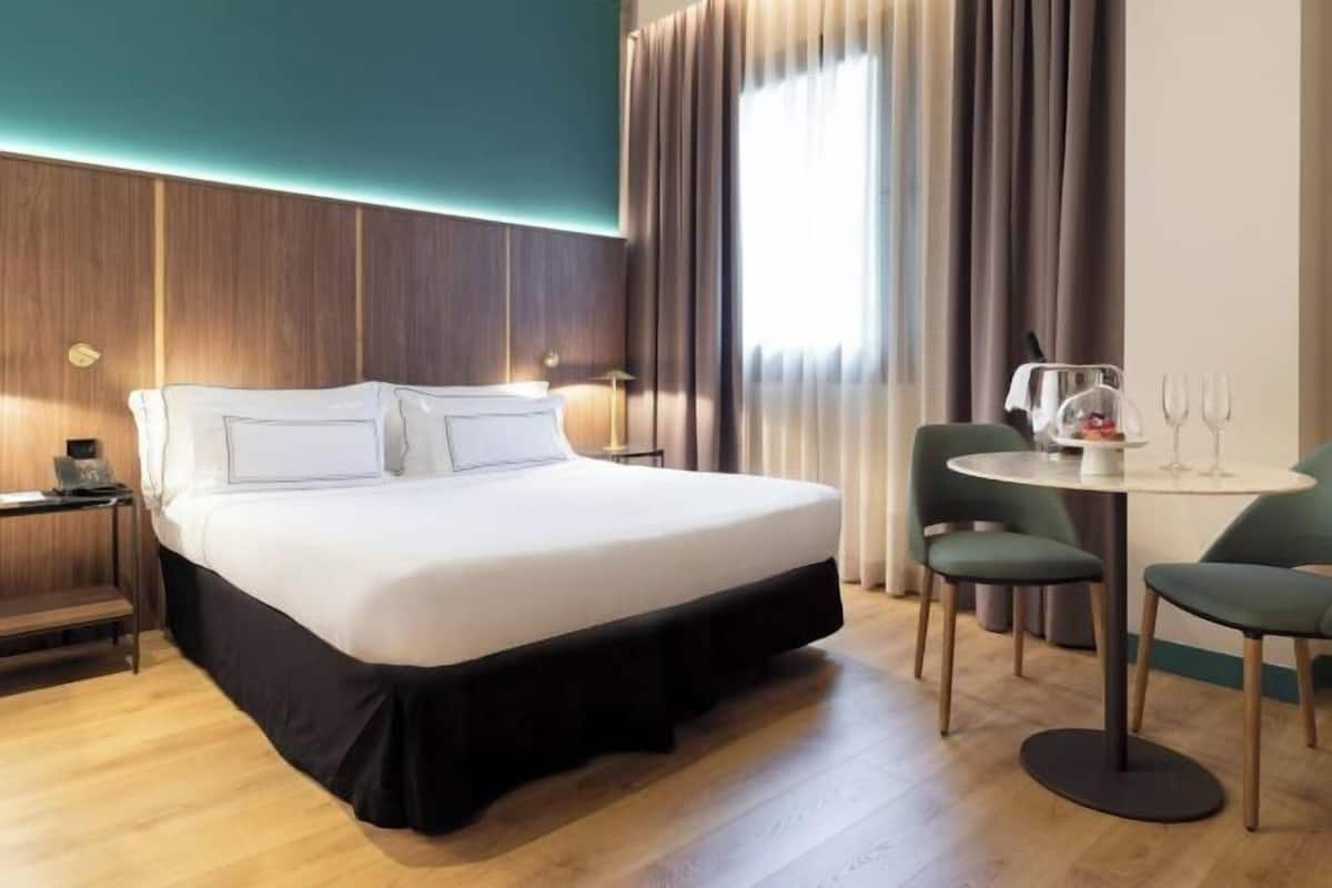Une chambre d’hôtel comprenant un lit, une table de chevet, une lampe, des rideaux, une fenêtre et un petit coin repas avec des chaises et une table.