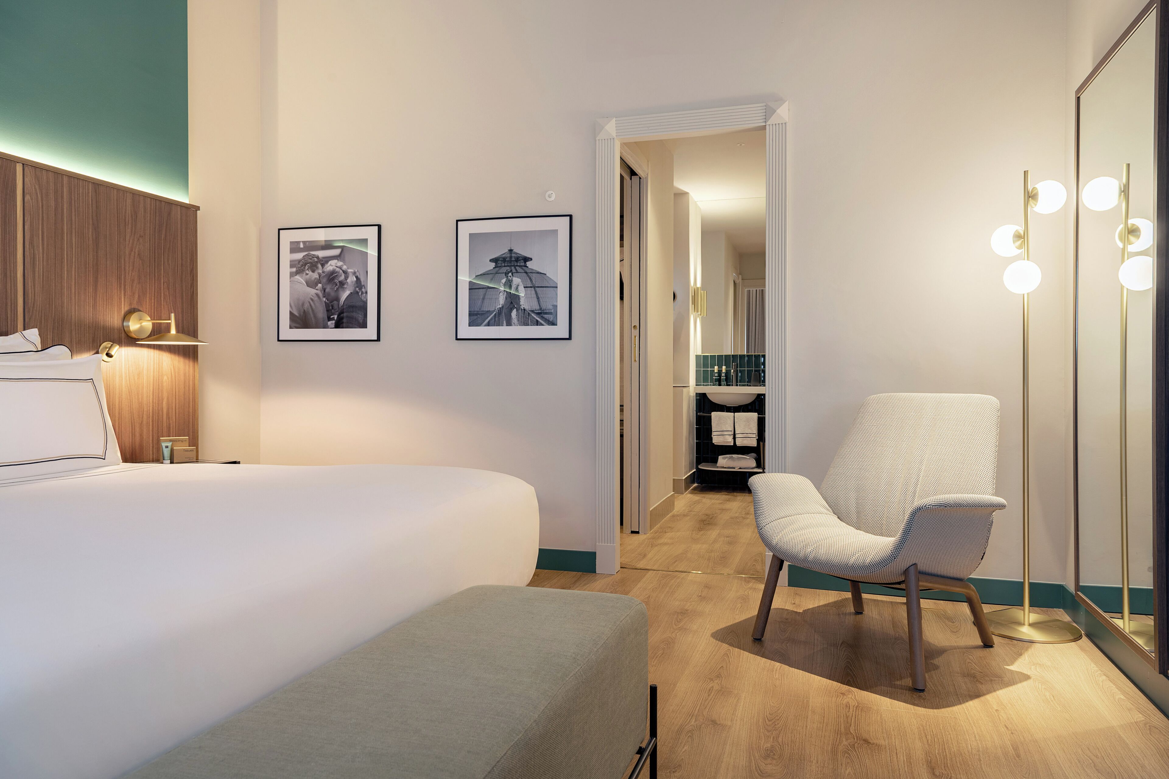 A modern hotel room with a large bed, a sitting area with a chair, a mirror with lights, and two framed pictures on the wall.