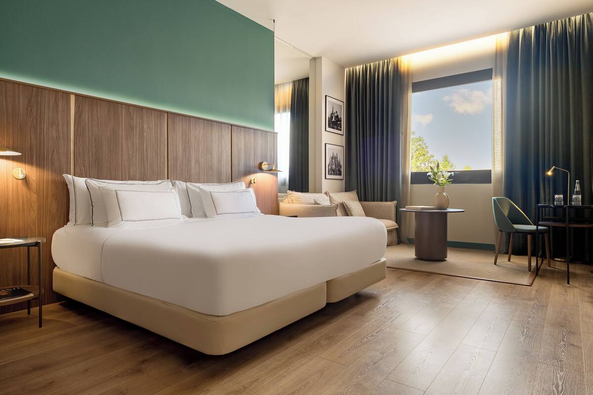 A modern hotel room with a large bed, a desk, a chair, a vase with flowers, and a view of the sky.