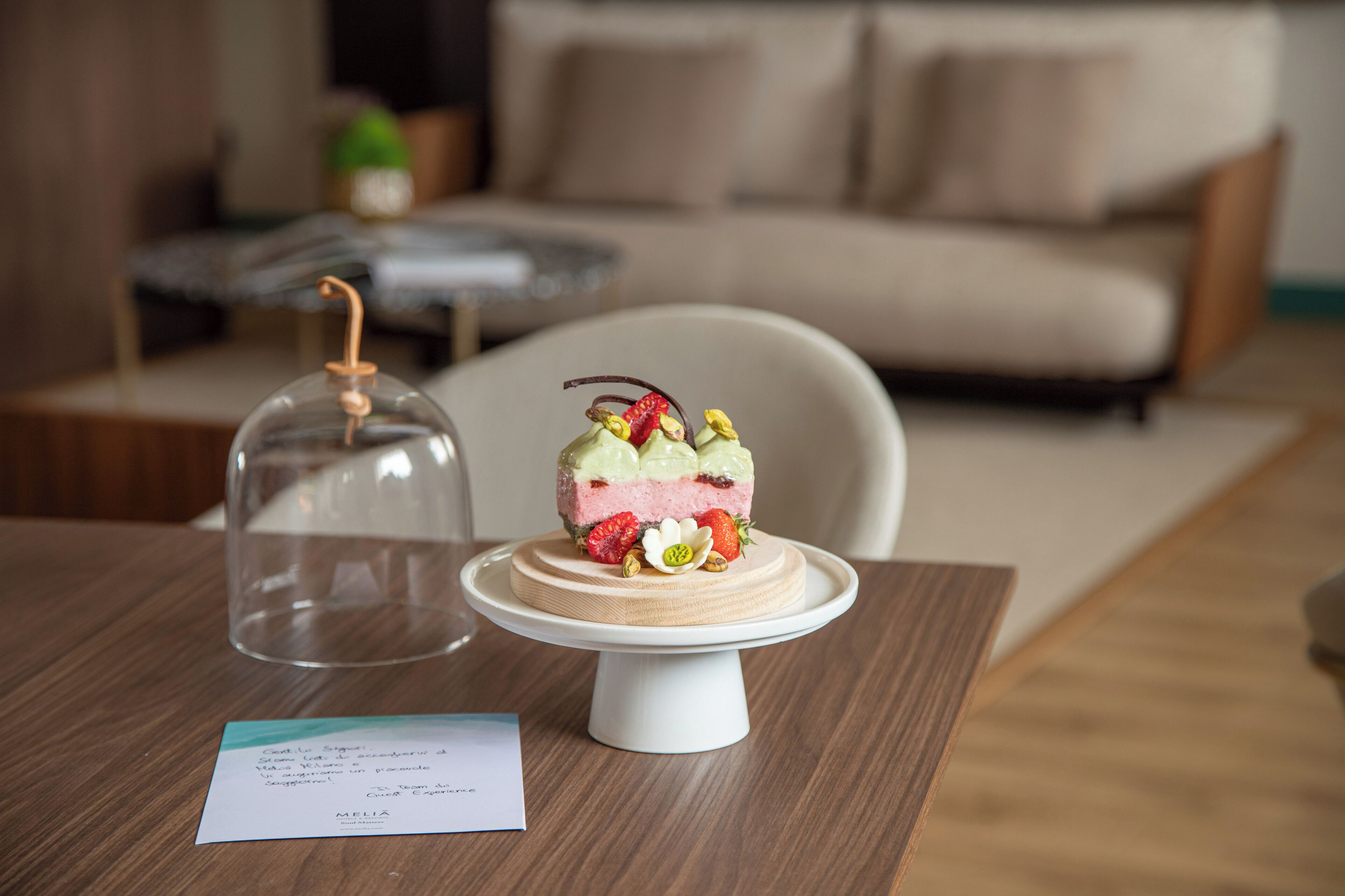 Un postre sobre un pedestal blanco con una campana de cristal, una tarjeta y una mesa de madera en un salón con un sofá y una mesa de centro.