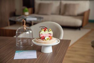 Un dessert su un vassoio bianco per dolci, sormontato da una cupola di vetro, con un biglietto e un tavolo di legno in un soggiorno arredato con divano e tavolino.