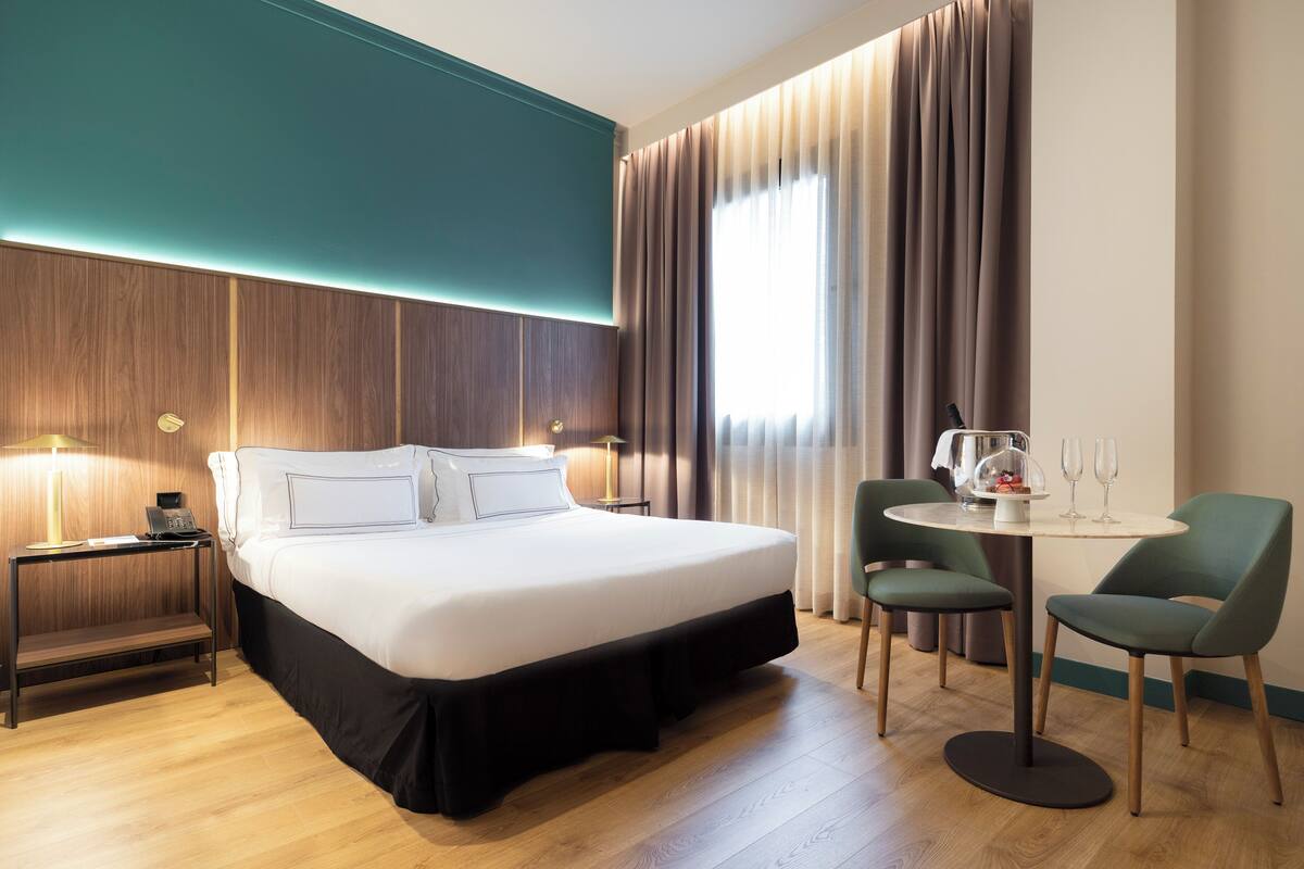 Une chambre d’hôtel avec un grand lit, des tables de chevet, une petite table ronde avec des chaises et une fenêtre avec des rideaux.