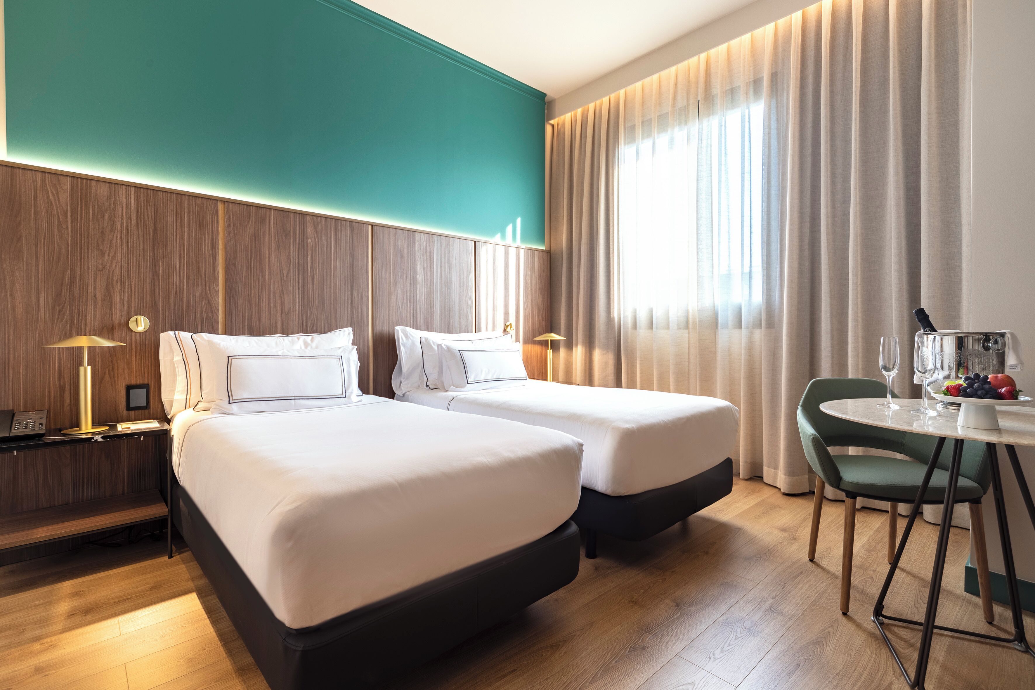 A hotel room with two beds, a small table with a fruit bowl, a chair, and a nightstand with a lamp.