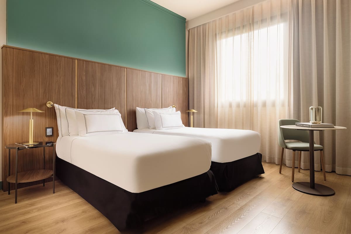 Une chambre d’hôtel avec deux lits, une petite table, une chaise et une fenêtre avec des rideaux.