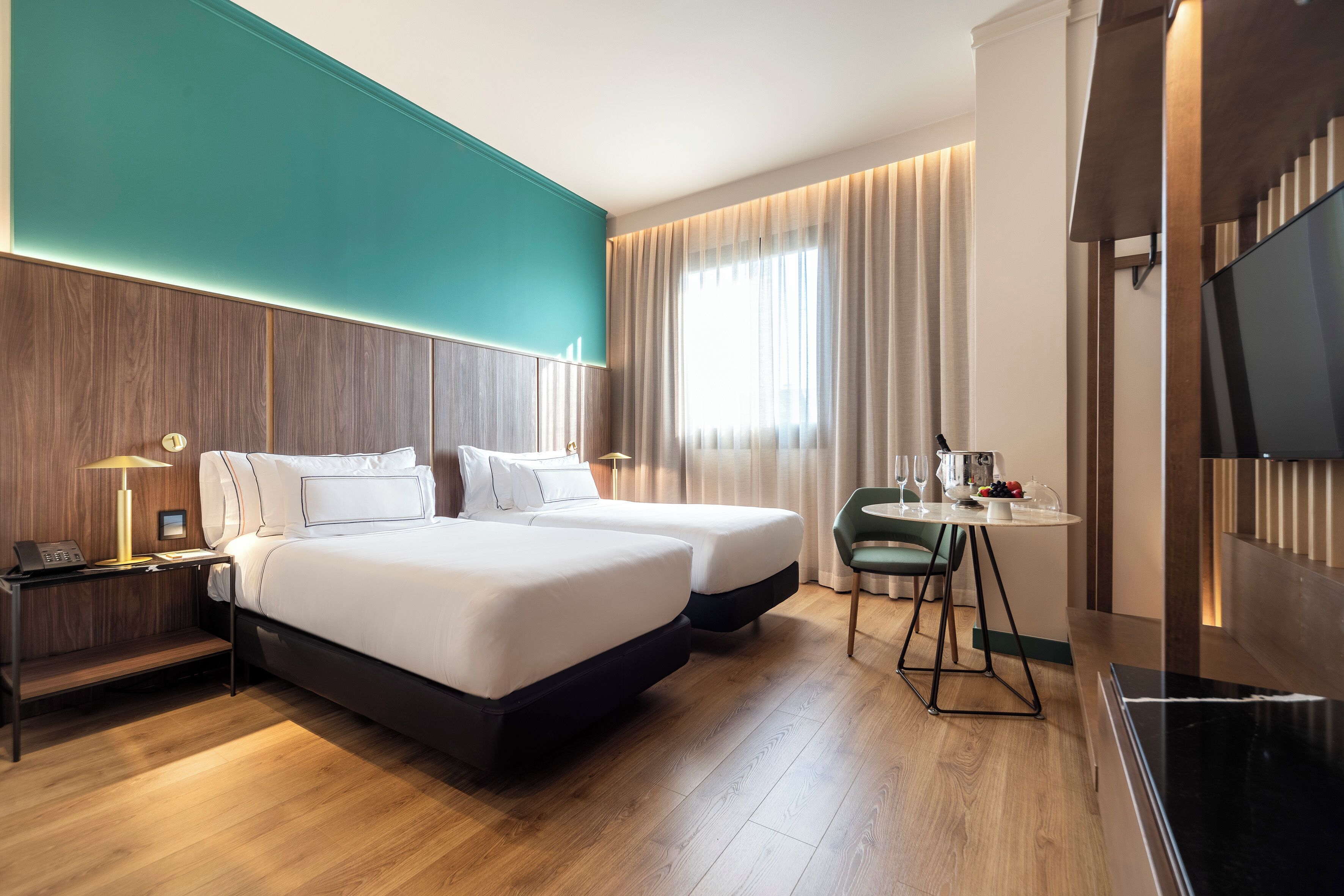 A hotel room with two beds, a desk, a chair, a TV, and a window with curtains.