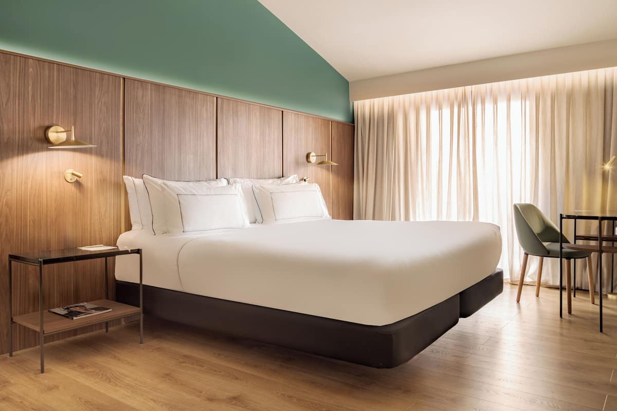 Une chambre d’hôtel moderne, dotée d’un grand lit, d’une table de chevet avec un magazine, d’un bureau avec une chaise et offrant une vue sur l’extérieur à travers des rideaux.