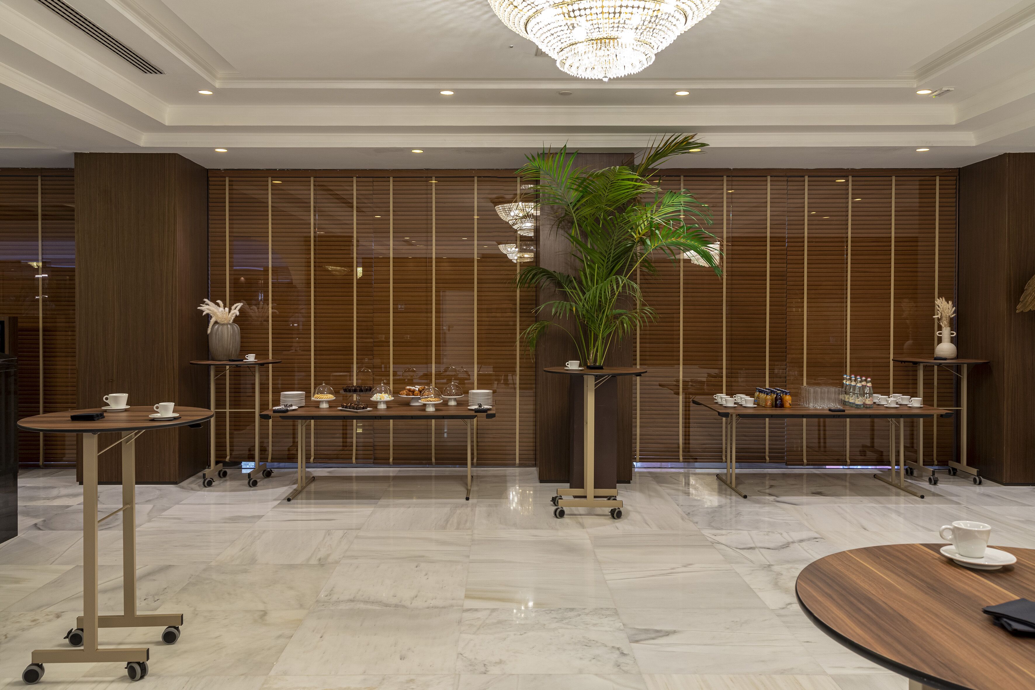Eine moderne Lobby mit Marmorboden, Holzvertäfelung und einer großen Kronleuchter. Mehrere Tische mit Teeservice und einer Topfpflanze sind vorhanden.