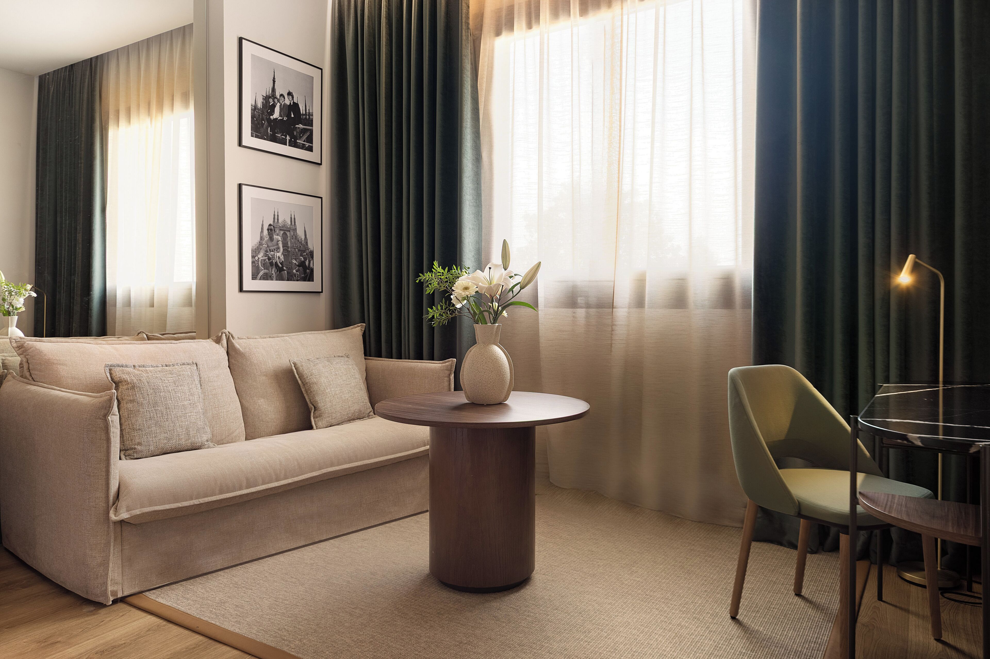 A modern living room with a beige sofa, a round wooden coffee table, a vase with flowers, a green chair, and a lamp.
